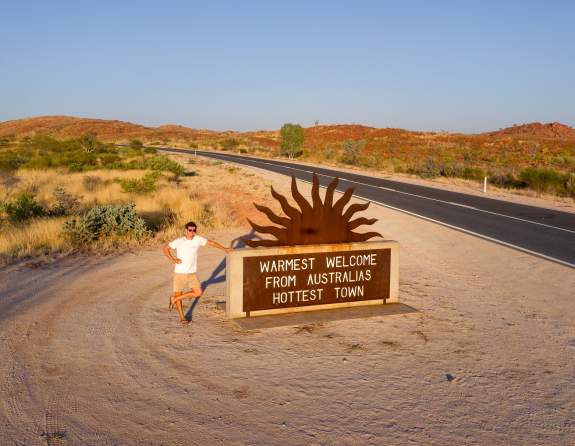 Man standing at the Marble Bar welcome sign which reads "Warmest welcome from Australia's hottest town."