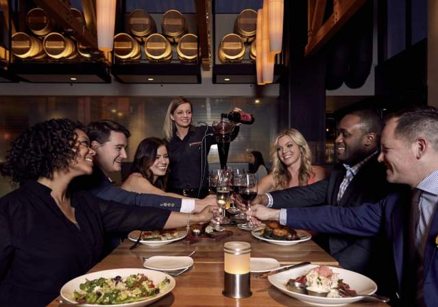 A group of six people toasting with glasses at a stylish restaurant table, featuring gourmet dishes and modern decor. Warm lighting enhances the inviting atmosphere.