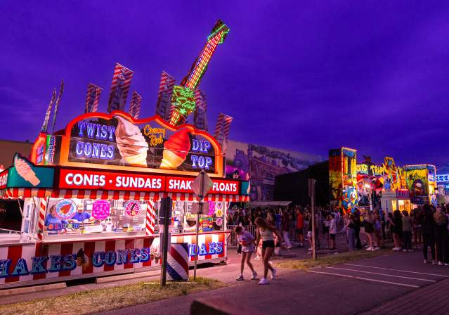 Brightly lit food stalls at a lively fair, featuring ice cream cones and sundaes, with a vibrant purple sky and crowds enjoying the atmosphere at Viva! Vienna!