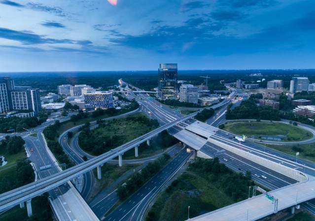 Aerial view of a busy highway interchange surrounded by modern buildings and greenery under a twilight sky in Tysons Corner. Traffic flows along multiple lanes.