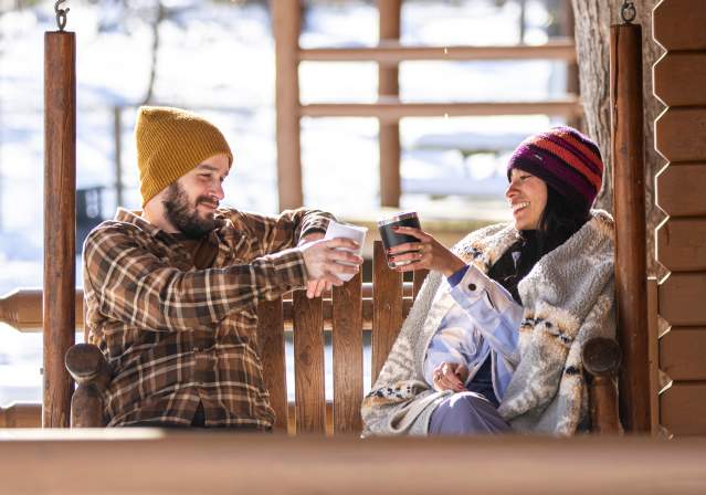 A couple at a cabin toasting with hot beverages while sitting on a wooden swing.