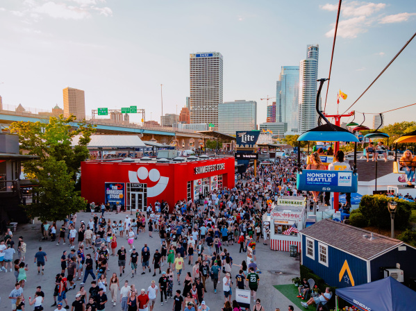A large crowd moves through the bustling Summerfest grounds in Milwaukee during the day, with the downtown skyline visible in the background. A red building with the Summerfest smiley-face logo serves as the festival store. Above, colorful gondolas on the Skyglider transport people across the venue. The scene is filled with booths, food stands, and signage under a clear sky.