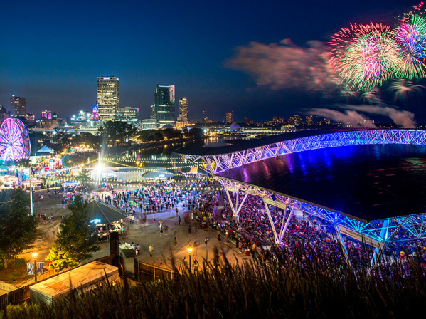A vibrant nighttime scene at Summerfest grounds in Milwaukee, featuring a large crowd, illuminated Ferris wheel, and the glowing roof of the American Family Insurance Amphitheater. The downtown skyline sparkles in the background as colorful fireworks burst over Lake Michigan, adding to the festive atmosphere.