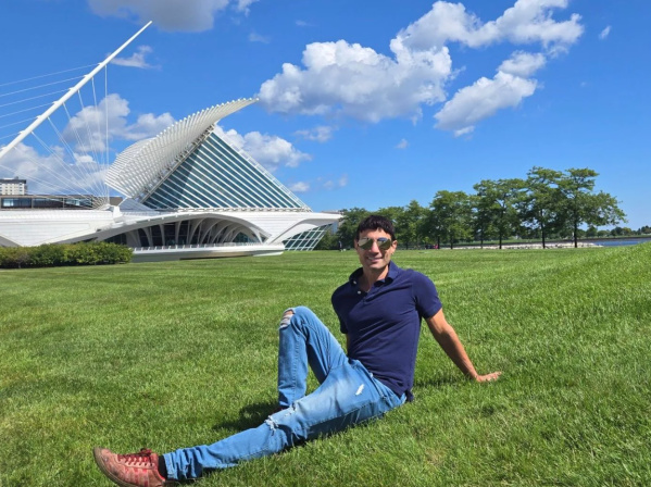 Person sitting relaxed on a grassy lawn in front of the Milwaukee Art Museum on a sunny day with blue skies and scattered clouds.