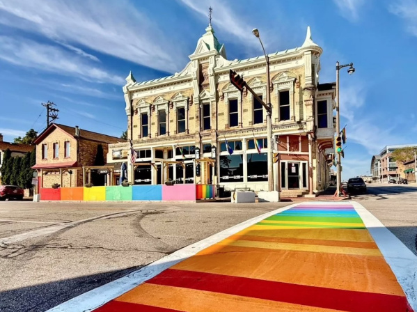 Historic cream-colored brick building with ornate architecture and rainbow pride flags, situated at a street corner with a brightly painted rainbow crosswalk leading toward it under a clear blue sky.
