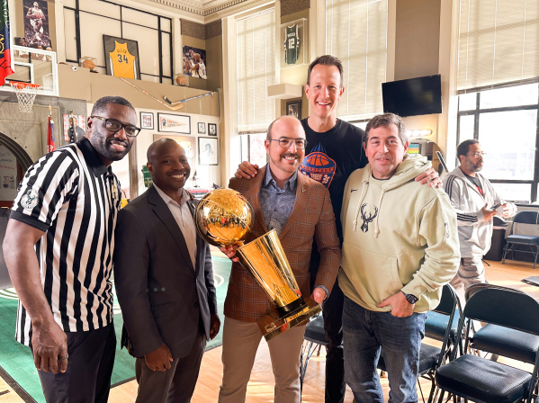 A group of five men pose together at Gee's Clippers. From left to right: Gee in a black and white striped referee shirt, Mayor Johnson in a dark business suit, David Caruso in a brown plaid blazer holding a golden basketball championship trophy,  Steve Novak in a dark shirt, and Peter Feigin in a cream-colored hoodie with a deer logo. The room features basketball memorabilia on the walls including framed jerseys and photos, a basketball hoop, and large windows.