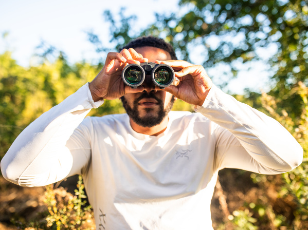 A man in a white long-sleeved shirt holds binoculars up to his eyes while standing outdoors in a sunny, tree-filled setting with green foliage and dappled sunlight in the background.