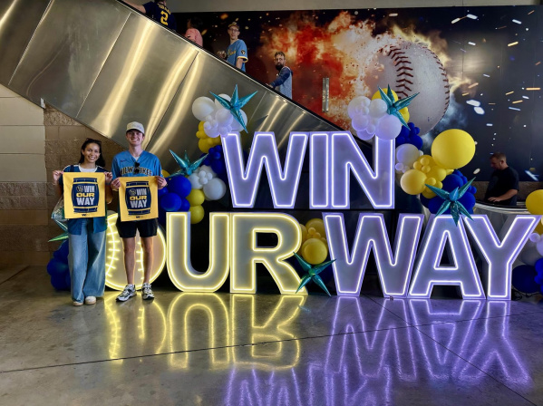 Two Milwaukee Brewers fans holding rally flags in front of a 'Win Our Way' sign.