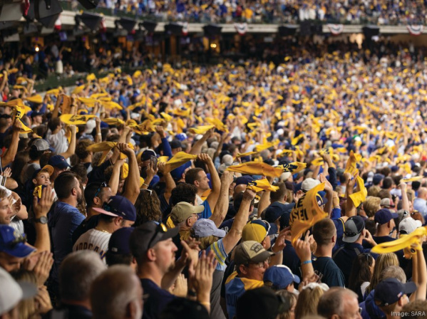 A packed stadium crowd cheers and waves yellow rally towels during a baseball game, filling the stands with motion and team colors.