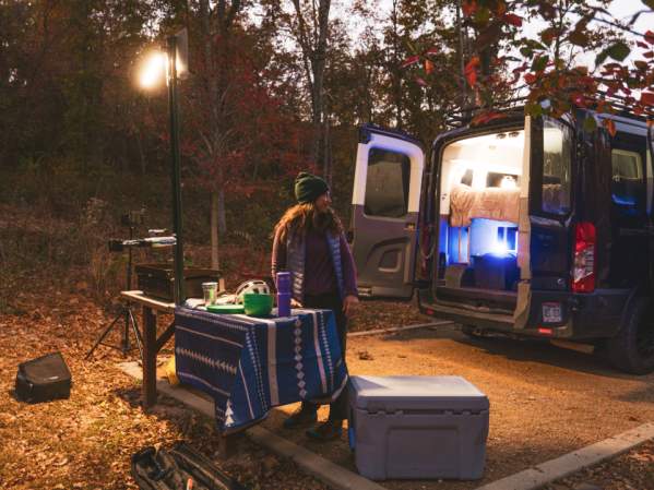 Person standing by a picnic table with camping gear and a lantern, next to an open van set up for sleeping in a wooded area at dusk.