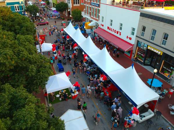 An aerial view of the Bentonville Square with green trees in the foreground and pedestrians strolling among vendor tents.