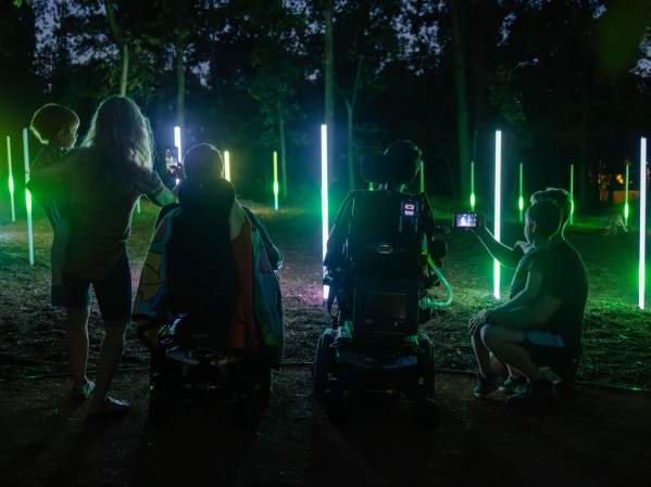 A family enjoys a vibrant light installation in a wooded area at night, with glowing green and white vertical lights illuminating the scene.