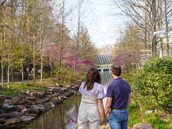 A couple holds hands while admiring the view over a rocky stream at Crystal Bridges Museum of American Art, surrounded by trees showing the colors of spring.