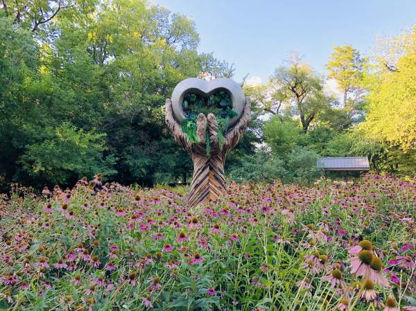 A large sculpture with hands that hold a heart. Pink flowers surround the sculpture.