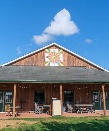 Barn on sunny day, barn quilt on top of barn and tables with chairs on patio.