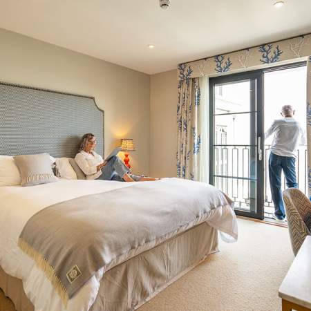 A man and a woman in a hotel room. Woman reading on the bed, man looking out of the window.