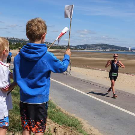 Two young boys cheering on their mum running along the race course waving welsh flags