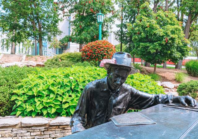 Hoagy Carmichael statue at Indiana University in Bloomington, Indiana, featuring a bronze figure seated at a piano in a landscaped garden with trees, shrubs, and a walkway in the background.