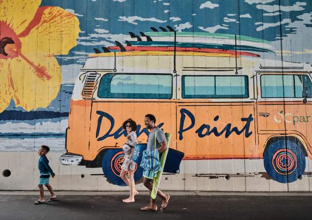 A family walks alongside a vibrant Dana Point mural featuring a van with surfboards on top.