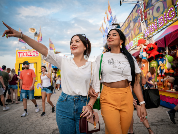 Two young women walk arm-in-arm through the Wisconsin State Fair, with one pointing excitedly ahead. Carnival rides, game booths, and crowds create a festive atmosphere in the background.