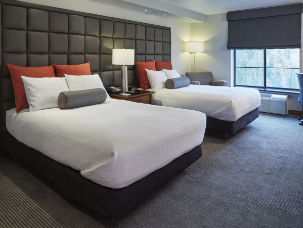 Modern hotel room with two queen beds featuring white bedding, dark upholstered headboards, bedside lamps, accent pillows, and a work desk near a window.