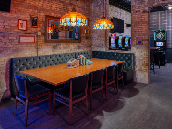 A cozy seating area featuring a long wooden table surrounded by dark chairs and a tufted green banquette. Warm stained-glass pendant lamps hang above the table, illuminating exposed brick walls decorated with framed art. In the background are bar games, including dartboards and illuminated gaming machines, creating a relaxed lounge atmosphere.