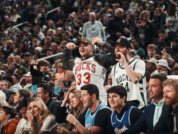Two excited fans wearing Milwaukee Bucks jerseys cheer from their seats during a packed game at Fiserv Forum, surrounded by a crowd of fellow spectators watching the action.