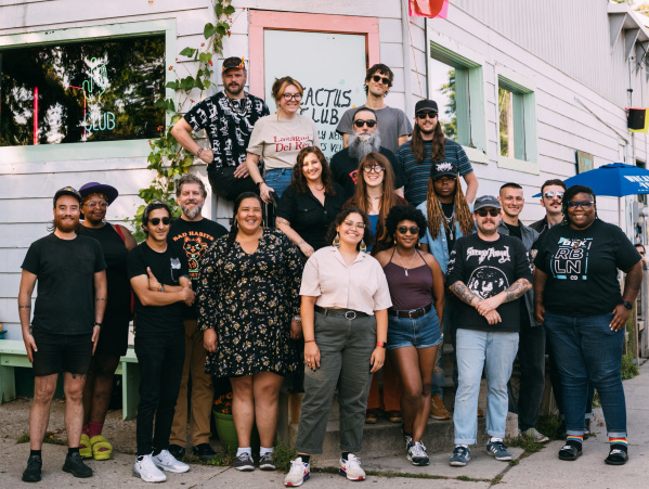 A large group of people stands smiling outside a light-colored building with a “Cactus Club” sign. They pose together on the sidewalk and steps, creating a friendly, community-focused group portrait.
