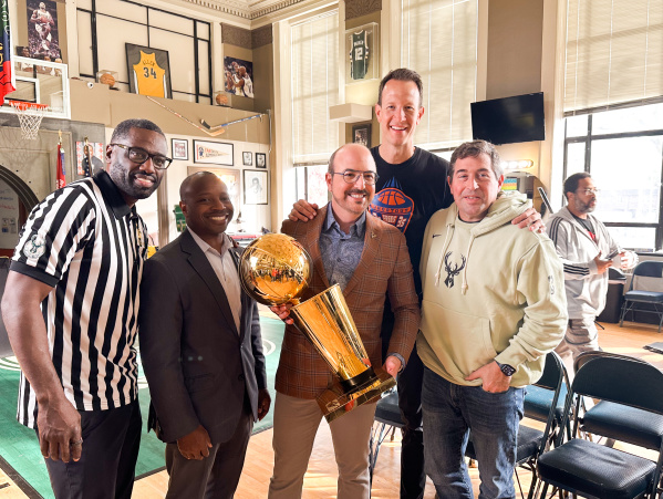 A group of five men pose together at Gee's Clippers. From left to right: Gee in a black and white striped referee shirt, Mayor Johnson in a dark business suit, David Caruso in a brown plaid blazer holding a golden basketball championship trophy,  Steve Novak in a dark shirt, and Peter Feigin in a cream-colored hoodie with a deer logo. The room features basketball memorabilia on the walls including framed jerseys and photos, a basketball hoop, and large windows.