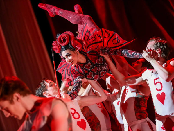 A performer dressed as the Queen of Hearts, wearing an elaborate red and black costume with heart patterns and curled horns, is lifted dramatically by dancers dressed as playing cards. The scene is bathed in red lighting, creating a bold and theatrical atmosphere reminiscent of Alice in Wonderland.