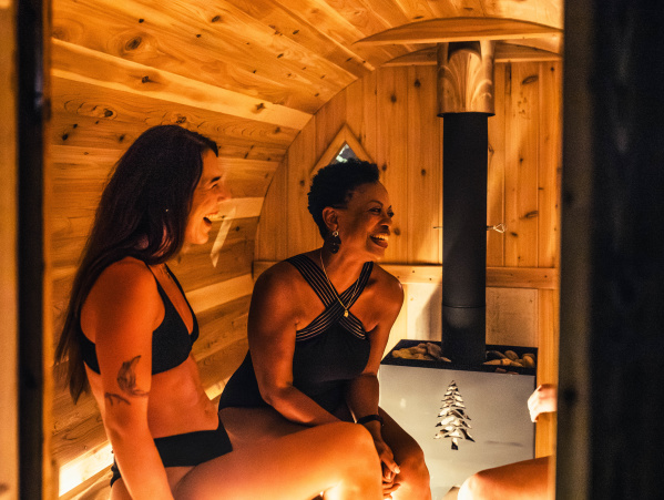 Two women in swimwear sit inside a warm, wood-paneled sauna at Heat Haven in Wauwatosa, smiling and talking near a small heater that glows softly in the center of the room.