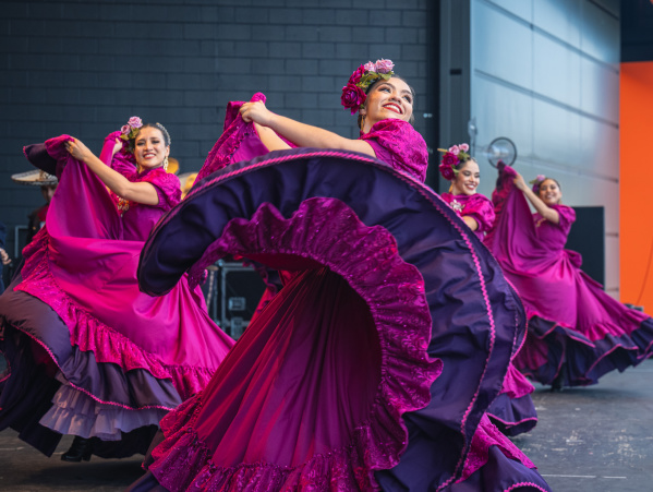 A group of female dancers performs a traditional Mexican folklórico dance on stage. They wear vibrant magenta dresses with purple ruffles and floral headpieces. The lead dancer in the foreground smiles brightly while gracefully twirling her flowing skirt. The dancers behind her mirror the movement, creating a colorful, energetic scene against a modern stage backdrop.