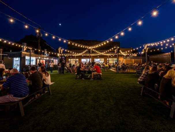 Image is of Covington Yard at night with twinkle lights above, people sitting at table and food trucks (operating out of containers) is lining the area.