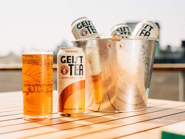 Several cans of Geist Tea sit in a metal bucket. One can sits on the table with a full glass next to it.