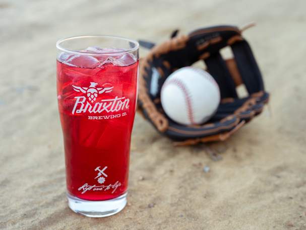 A glass with the Braxton Brewing logo sits in the sand next to a baseball glove. The glass is full of Rally, Braxton's cranberry hard seltzer
