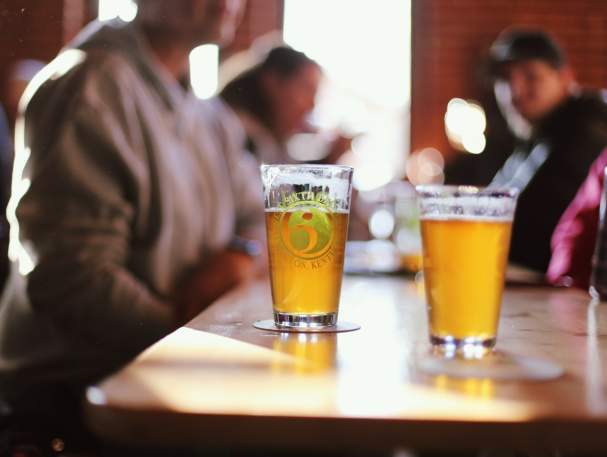 Focused on two glasses of beer sitting on a table. There are out of focus people in the background.