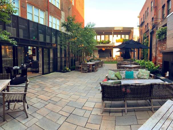 An outdoor patio, lush with plants, offers seating. A fireplace is to the right and an area that can be used as a stage is in the background.