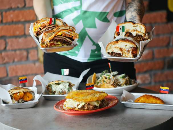 A person offering the viewer tortas in both hands with a table of tortas, empanadas and more food below