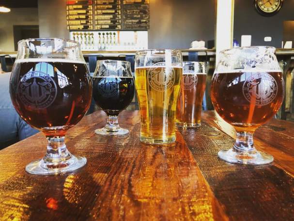 five glasses of different colored beer on a wooden table with a menu board in the background at wooden cask brewery in newport kentucky