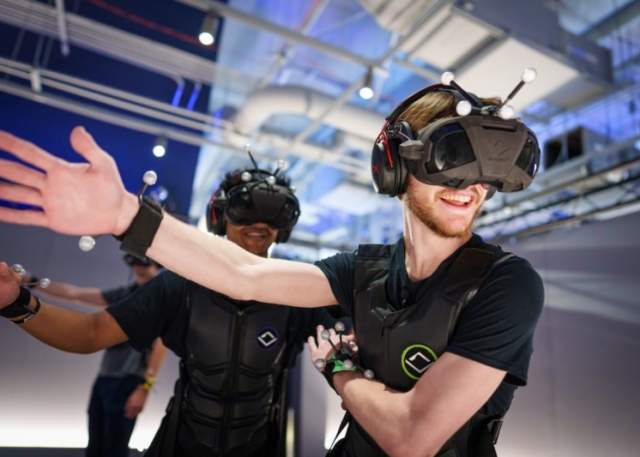 Two men wearing virtual reality headsets and motion sensors extend their arms at Sandbox VR