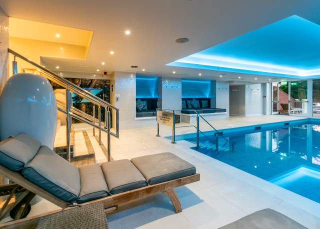Indoor pool area with modern design features, such as a blue-lit ceiling and sleek lighting. There are cushioned lounge chairs and glass doors leading outside, creating a serene ambience.