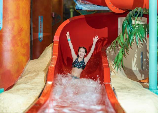 A young girl sliding down a water slide with her arms in the air in an indoor waterpark