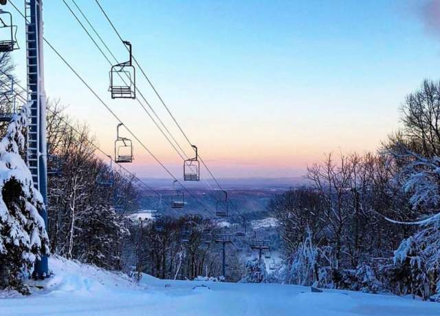 Sunset view from a ski mountain with trees along either side of the ski slope and a ski lift in frame.