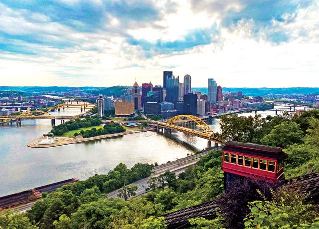 An aerial view of a red cable car moving down a side of a mountain with a skyline of Pittsburgh in the background