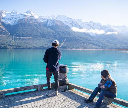 A man fishing off a wharf over a vibrant blue lake and snow covered mountains, while another sits and watches.