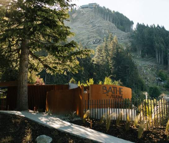 Entrance to a rustic outdoor sauna retreat surrounded by native forest and alpine hillside landscape, featuring timber fencing and natural materials in a secluded setting.