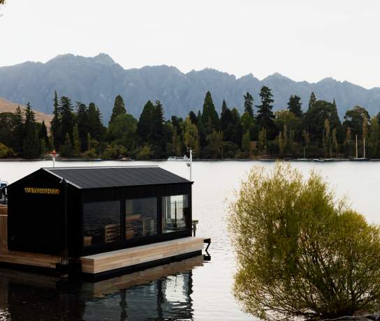 Modern floating sauna building on a lakeside dock, surrounded by calm water, alpine mountains, and autumn trees in a peaceful natural setting.