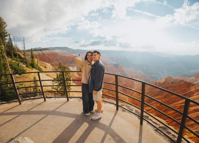 A couple stands at a paved scenic overlook at sunset, taking a selfie with the bright orange and red rock formations of Cedar Breaks National Monument behind them.