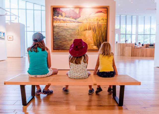 Three kids sitting on a wood bench in front of a landscape painting at the Southern Utah Museum of Art.