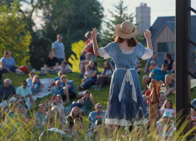 Actor in straw hat and dress faces a gathered crowd on the Green of the Beverley Center for the Arts for the Utah Shakespeare Festival Greenshow.
